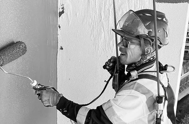 Alpine-Tech 360 rope access technician painting a building wall with a roller while suspended on ropes