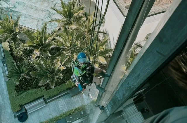 Alpine-Tech 360 rope access technician in a blue helmet spraying water onto a glazed facade for a leak test, seen from above a landscaped courtyard