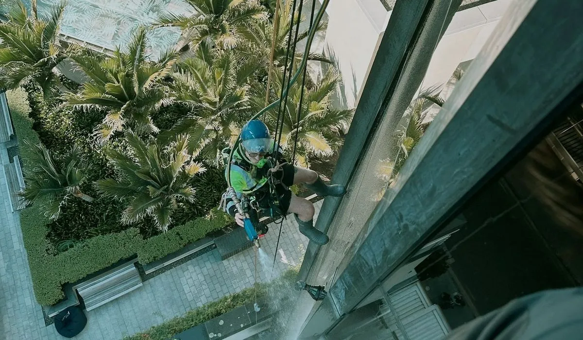 Alpine-Tech 360 rope access technician in a blue helmet spraying water onto a glazed facade for a leak test, seen from above a landscaped courtyard