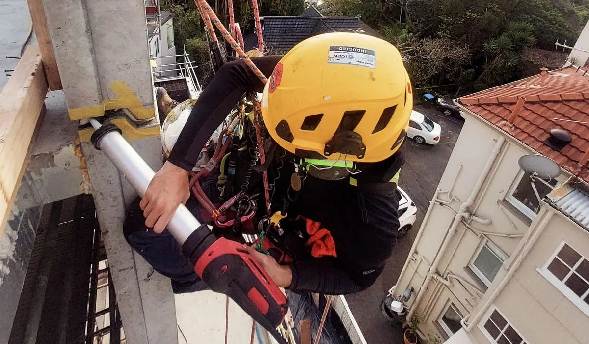 Alpine-Tech 360 rope access technician in a yellow helmet sealing a facade joint with a caulking gun, high above the Auckland skyline
