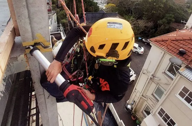 Alpine-Tech 360 rope access technician in a yellow helmet sealing a facade joint with a caulking gun, high above the Auckland skyline