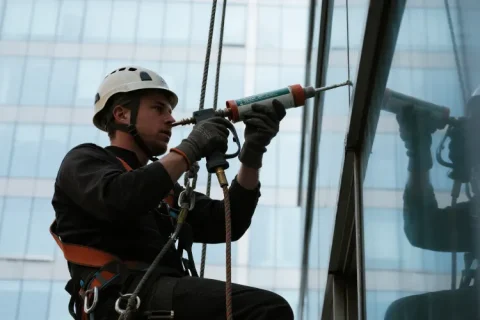 A rope access worker sealing window joints on a high-rise façade with a caulking gun, wearing a helmet, gloves, and harness.