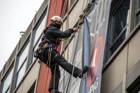 Rope access technician installing a large banner sign on a tall building, securing it with ropes, wearing helmet and harness.