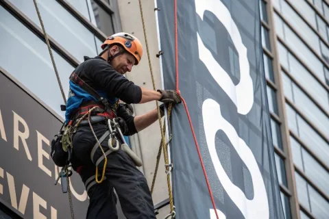 Rope access technician installing a large banner sign on a tall building, securing it with ropes, wearing helmet and harness.