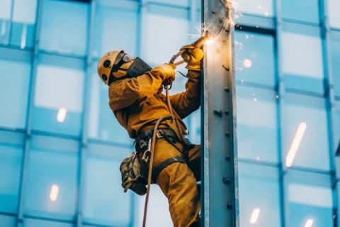 Rope access welder repairing a steel beam on a tall building façade, sparks flying from welding torch, wearing helmet, harness, gloves, and welding mask.