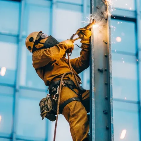 Rope access welder repairing a steel beam on a tall building façade, sparks flying from welding torch, wearing helmet, harness, gloves, and welding mask.