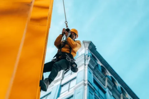 Rope access inspector photographing and documenting high-rise façade condition, DSLR camera in hand, wearing safety harness and helmet.